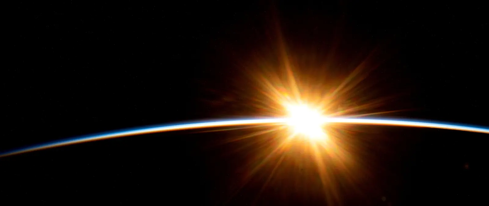 Sunrise breaking over Earth's horizon, seen from orbit: a thin blue band of atmosphere against black.