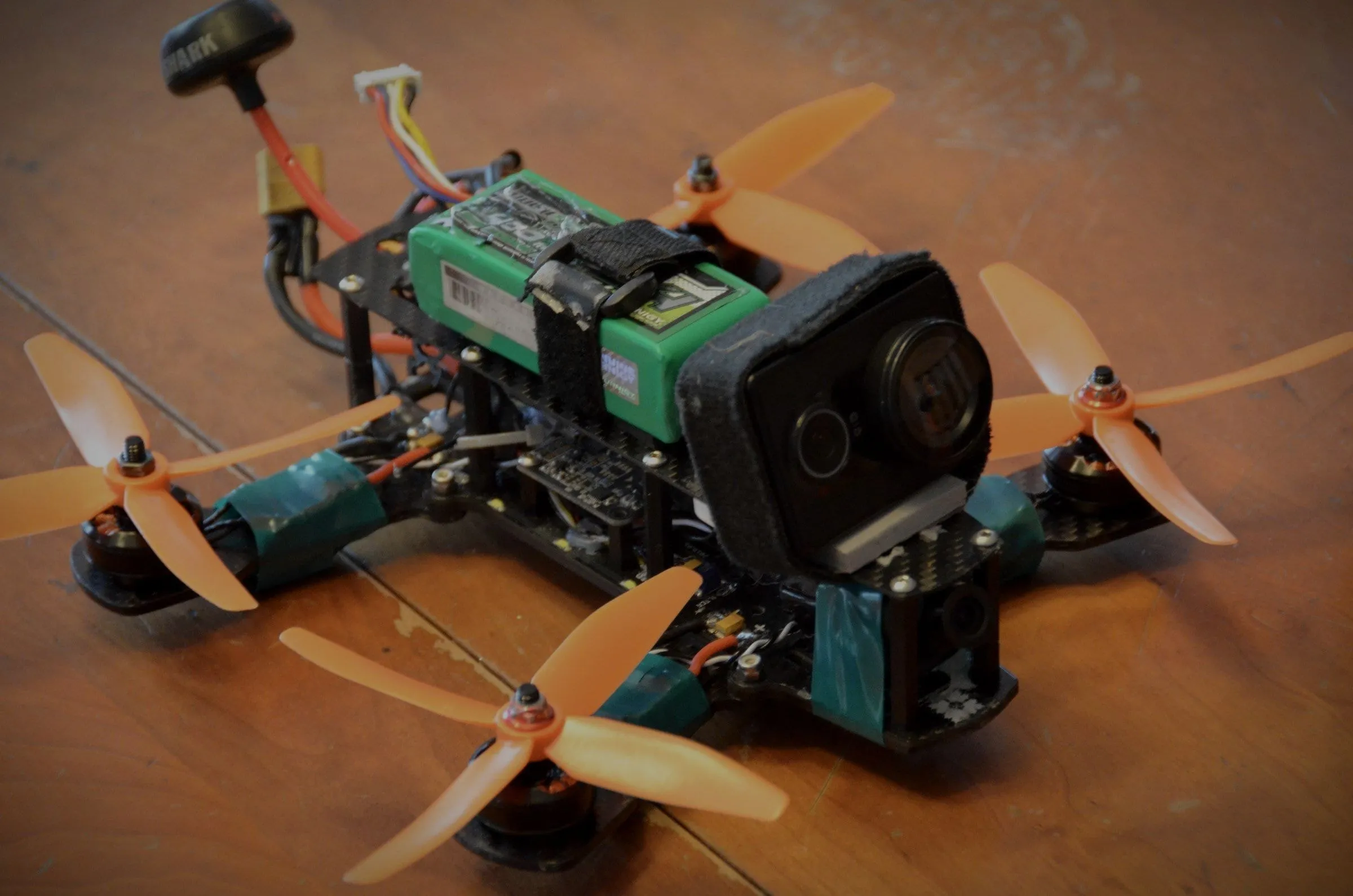 A home-built FPV racing quadcopter on a workbench: carbon frame, orange props, strapped battery, action camera.